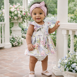 Baby in a floral dress standing on a porch with flowers in the background