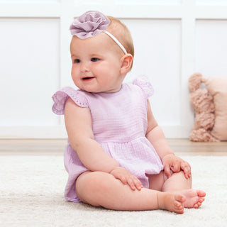 Baby in a purple outfit with a flower headband sitting on a white surface.