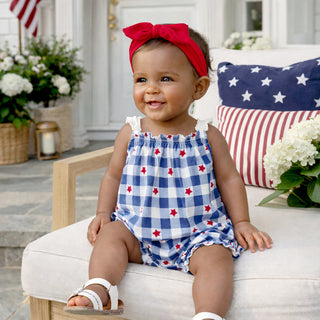 Baby in a blue checkered romper with red stars sitting on a chair outdoors.