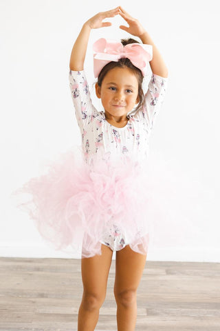 Young girl in a ballerina print tutu leotard with a large pink bow standing on a wooden floor.