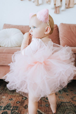 Young baby girl in a pink tutu leotard standing by a couch with decorative pillows.