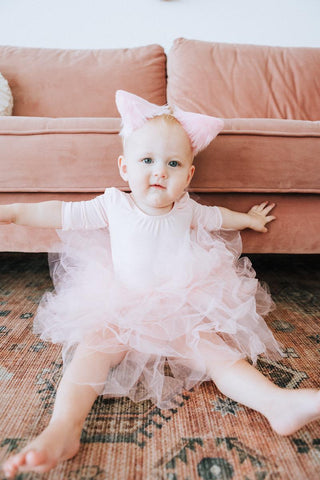 Baby in a pink dress with a large bow sitting on a patterned rug.