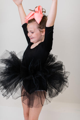 Young girl in a black ballet outfit with a pink bow and black tutu against a plain background
