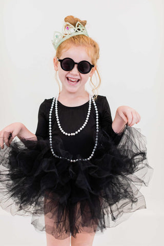 Young girl wearing a black leotard with a tutu, sunglasses, and a tiara on a white background
