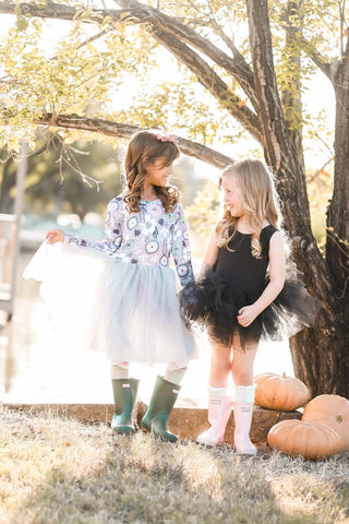 Two young girls in dresses standing outdoors with pumpkins and a tree in the background