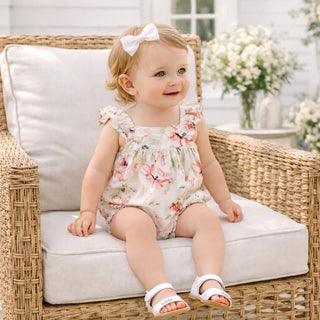 Baby girl in a floral romper sitting on a wicker chair with a white cushion.