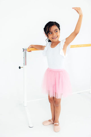 Young girl in a white leotard and pink skirt standing on one leg with arms raised, leaning against a ballet barre.