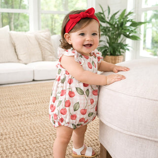 Baby in a cherry-patterned romper standing on a beige rug with a white couch and plant in the background.