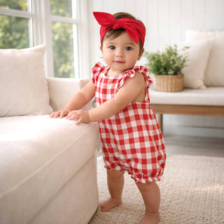 Baby in a red checkered outfit standing in a living room.