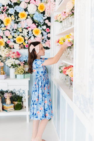 Young girl in a floral dress standing in front of a wall of flowers, reaching for a flower arrangement.