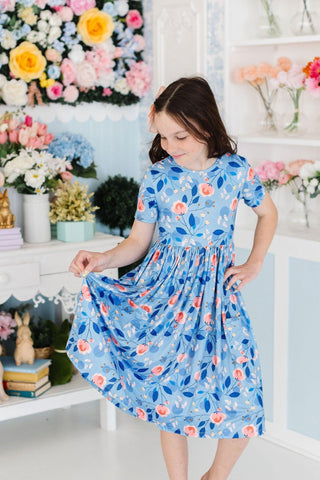Young girl in a blue floral dress standing in a room with decorative flowers and shelves.