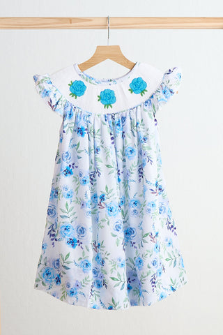 A hand-smocked dress with a floral print, predominantly blue with a white background, displayed on a wooden hanger.