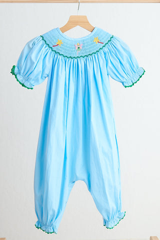 A blue girl's romper with hand smocked details, green trim, and a fairy theme embellishment on the collar and cuffs.