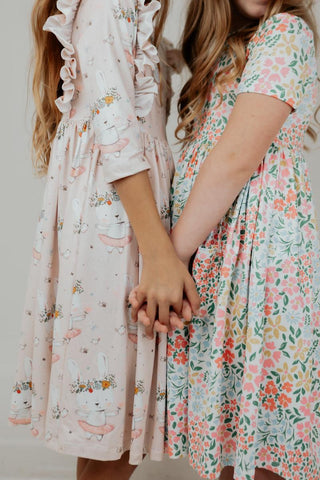 Two girls holding hands, wearing floral dresses against a neutral background