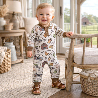 Baby wearing a farm patterned onesie standing in a room with wooden furniture and large windows.