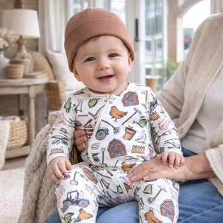 Baby wearing a farm patterned onesie and hat, sitting on a person's lap indoors.