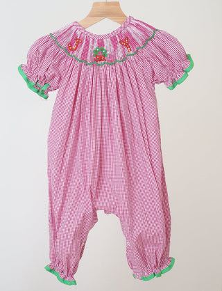 A pink gingham romper with green and red embroidery and a smocked waist, displayed on a wooden hanger.