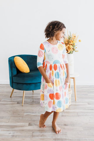 Young girl wearing a colorful dress standing in a room with a blue chair and yellow flowers.