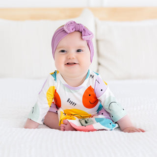 Baby wearing a colorful outfit with smiley prints on a white bed.