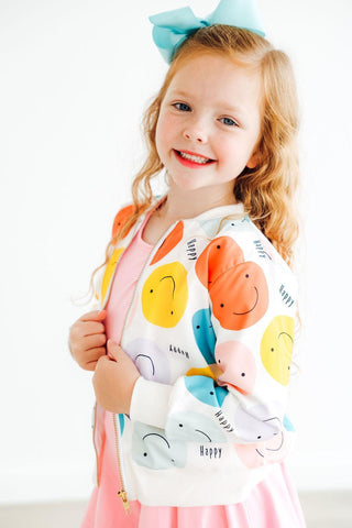 Young girl wearing a colorful jacket with smiley faces and a pink dress.