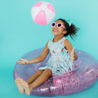 Child sitting on a pink inflatable ring with a beach ball, wearing sunglasses against a blue background