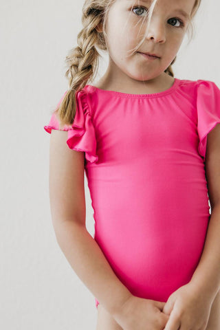 Young girl wearing a bright pink leotard with ruffled sleeves against a plain background
