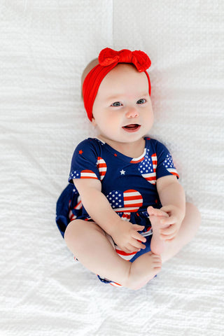Baby wearing a blue onesie with American flag design and a red headband on a white textured surface.
