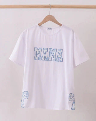 White t-shirt with 'MAMA' printed on it, hanging on a wooden hanger against a plain background.