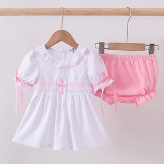 White dress with pink trim and matching bloomers on hangers against a white background