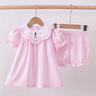 Pink baby outfit with a top and shorts on a hanger against a light pink background