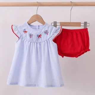 Baby outfit with ruffled neckline and red bloomers on a wooden rack against a light pink background