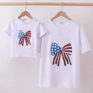 Two white t-shirts with sequin bow designs on a wooden rack against a white background