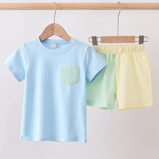 Children's light blue shirt with green pocket and yellow shorts hanging on a wooden rack against a white background.