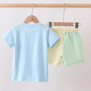 Children's pastel outfit with a blue shirt and green shorts hanging on a wooden rod.