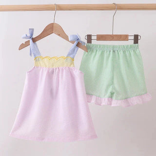 Children's pink top and green shorts with ruffles hanging on wooden hangers against a white background.