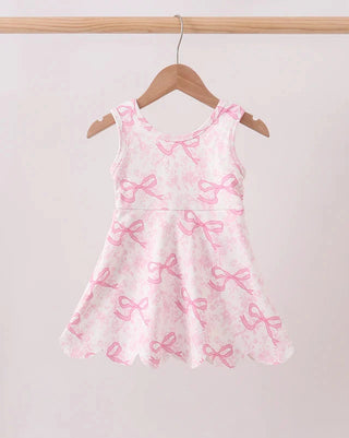 A sleeveless white dress with a pink bow pattern, hanging on a wooden hanger from a light wood rod against a plain light background.