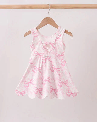 A sleeveless white dress with a pink bow pattern, hanging on a wooden hanger from a light wood clothing rack against a plain light background.