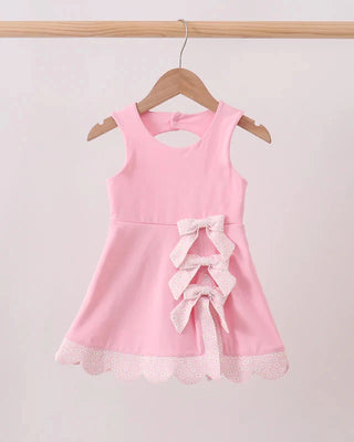 A pink sleeveless dress with a lace bow detail on the front, hanging on a wooden hanger against a light pink background.
