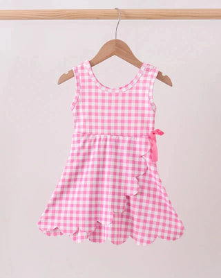 A pink and white gingham sleeveless dress for a toddler girl, hanging on a wooden hanger from a light wood rod against a plain light beige wall.
