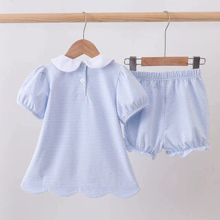 A baby girl's two-piece outfit set consisting of a light blue and white striped short-sleeve blouse with a white Peter Pan collar and scalloped hem, paired with matching light blue and white striped ruffled bloomers, hanging on wooden hangers against a plain light gray wall.