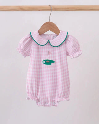 A pink and white gingham baby romper with a green-trimmed Peter Pan collar and a green golf ball graphic on the chest, hanging on a wooden hanger from a wooden clothes rack.