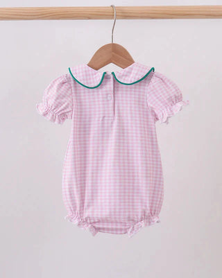 A pink and white gingham baby romper with a green-trimmed Peter Pan collar, hanging on a wooden hanger from a light wood clothes rack against a plain off-white wall.