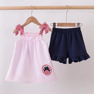 A toddler girl's two-piece clothing set hanging on wooden hangers against a plain light background. The set includes a light pink sleeveless dress with red-and-white gingham bow details on the shoulders and a matching circular patch on the lower left, paired with navy blue ruffled shorts.