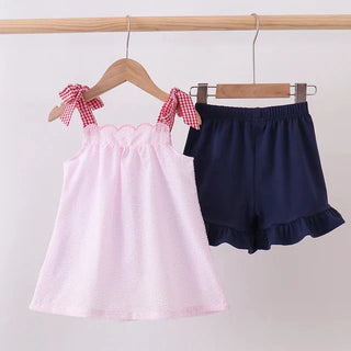 A two-piece girls' outfit set hanging on wooden hangers against a plain light background. The set includes a light pink sleeveless top with red-and-white gingham bow details on the shoulders and navy blue ruffled shorts.