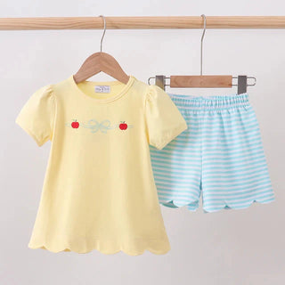 A children's two-piece outfit set hanging on wooden hangers against a plain light background. The set includes a pale yellow short-sleeve top with small red apple and bow embroidery on the chest, and light blue-and-white striped shorts with scalloped hems.