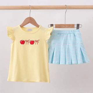 A two-piece girls' outfit hanging on wooden hangers from a wooden rod. The set includes a pale yellow short-sleeve top with a red apple graphic on the chest, and a light blue and white striped pleated mini skirt.