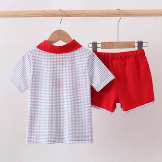 A children's two-piece clothing set hanging on wooden hangers from a light wood rod. The set includes a short-sleeve button-up shirt with a red collar and a white gingham pattern with thin red and blue lines, paired with solid bright red shorts featuring a white gingham trim.