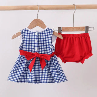 A baby girl's two-piece outfit hanging on wooden hangers from a wooden rod. The set includes a blue and white gingham check sleeveless dress with a red bow at the waist and a pair of bright red bloomers.