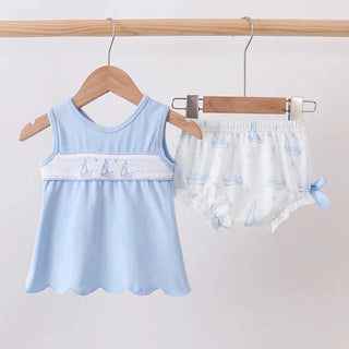 A baby clothing set hanging on wooden hangers from a wooden rod, featuring a light blue sleeveless top with a white embroidered band and scalloped hem, paired with white diaper cover pants printed with light blue sailboats.