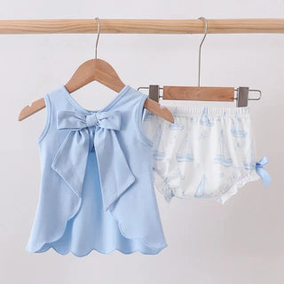 A baby girl's two-piece outfit hanging on wooden hangers from a wooden rod. The set includes a light blue sleeveless top with a large ruffled bow at the back and matching light blue ruffle trim along the hem, paired with white diaper cover shorts featuring a light blue sailboat pattern.